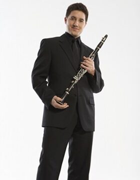 Man in black suit holding a clarinet, smiling.