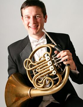 Man in tuxedo holding a French horn and smiling.