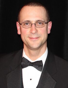 Man in tuxedo and glasses posing against a black backdrop.